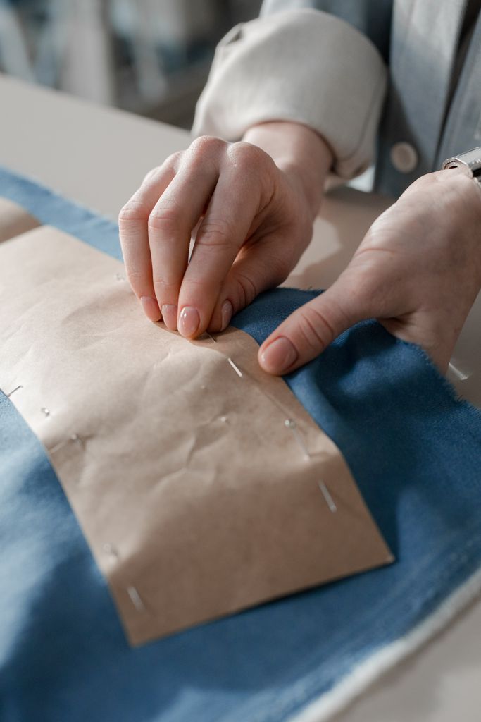 Detailed image of hands sewing a blue fabric with a pattern pinned in place, showcasing dressmaking process.